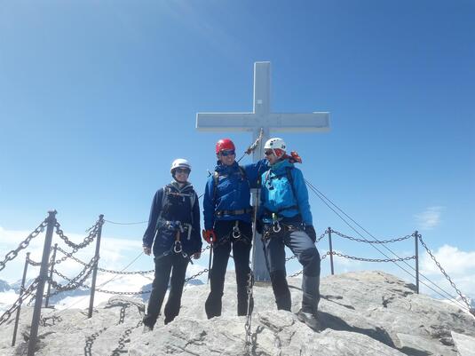 Three mountain climbers are standing on a summit next to a large cross. The surroundings are rocky and the sky is clear.