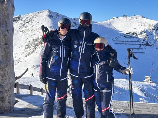 Drei Skifahrer stehen auf einem Berg im Schnee. Im Hintergrund sind schneebedeckte Hänge und blauer Himmel zu sehen.