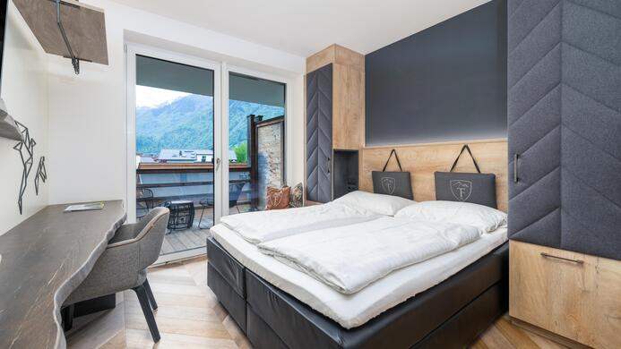 A modern bedroom with a double bed and wooden furniture. A small desk and a balcony overlooking the mountains are also visible.