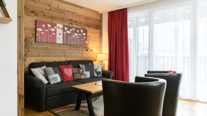 Ein gem&uuml;tliches Wohnzimmer mit einer Holzwand und einem Sofa. Es gibt einen Tisch und zwei schwarze Sessel, sowie einen gro&szlig;en Fensterbereich mit Tageslicht.