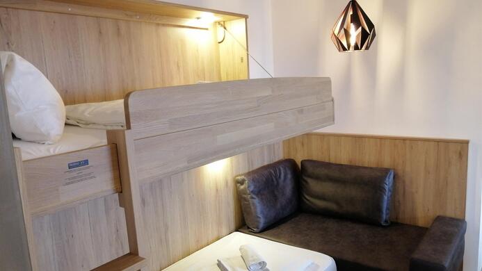 A modern bedroom with a loft bed and a cozy sitting area. The walls are light, and there is a stylish lamp.