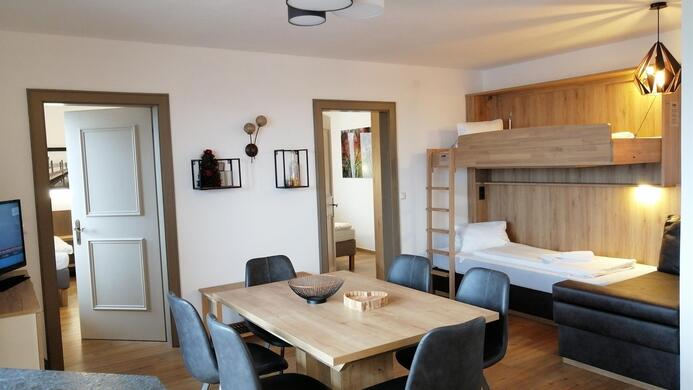 A bright, modern room with a cozy seating area and a dining table. There is a loft bed and a TV on the wall.