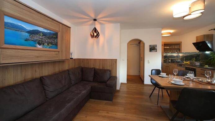 A modern living room with a large beige sofa and an elegant dining table. A picture of a coastal landscape hangs on the wall, and the atmosphere is bright and friendly.