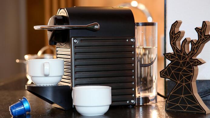 A modern coffee machine is on a countertop with two cups and a coffee capsule. In the background, there is a decorative figure in the shape of antlers.