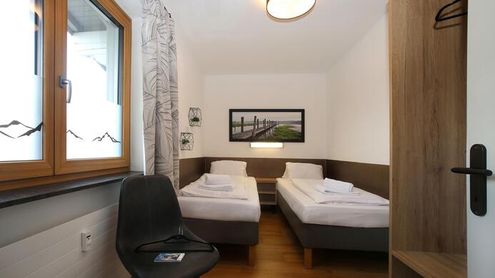 A simple room with two single beds, a chair, and a window. The walls are light-colored, and pleasant lighting creates an inviting atmosphere.