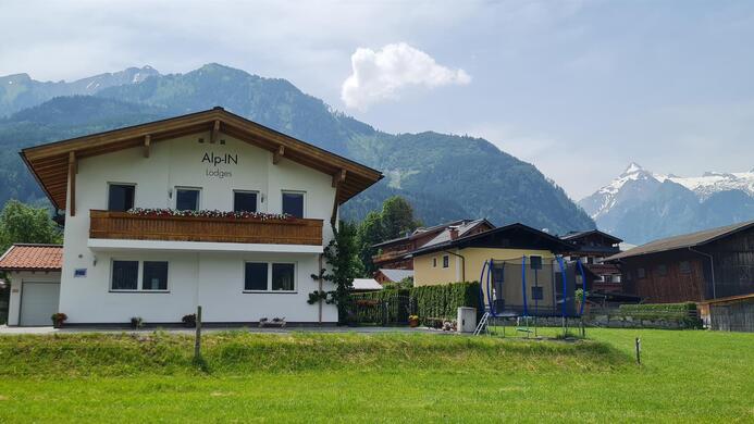 A charming house in a rural setting with mountains in the background. The scene shows a peaceful, green meadow and a clear blue sky.