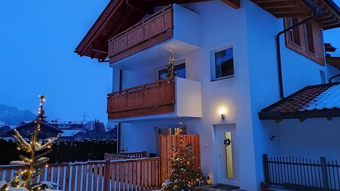 A modern house in winter with a snow cover. In front of the house stands a decorated Christmas tree and it is dusk.