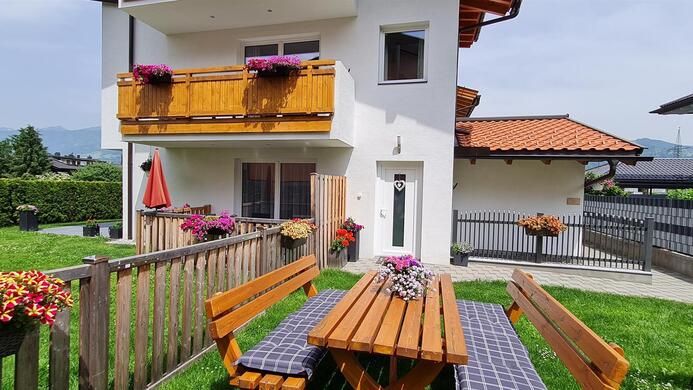 A modern house with a balcony and blooming plants. In the foreground, there is a wooden table with a bench in the garden.