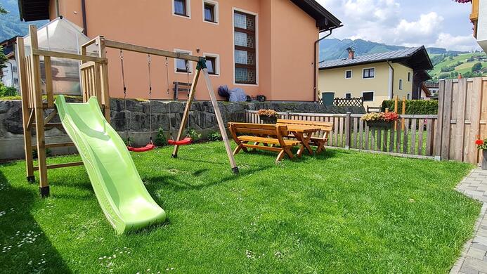 A cozy garden with a slide, a swing, and a wooden playground. In the background, modern houses and blooming flowers can be seen.