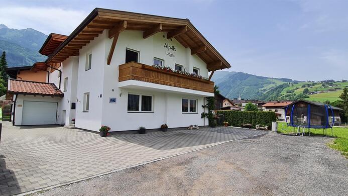 A modern house with a wooden balcony in a quiet setting. In the background, green hills and mountains can be seen.