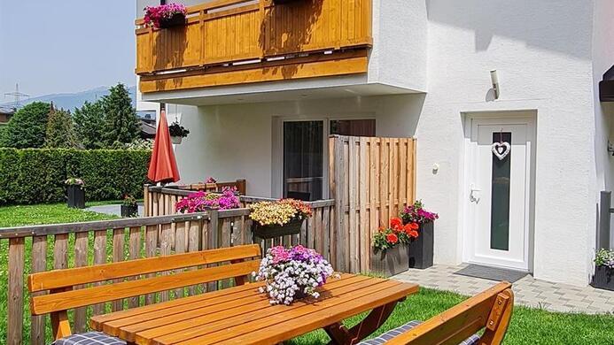 A cozy wooden terrace with tables and benches is situated on a green meadow. In the background, you can see a bright building with balconies and flowers.