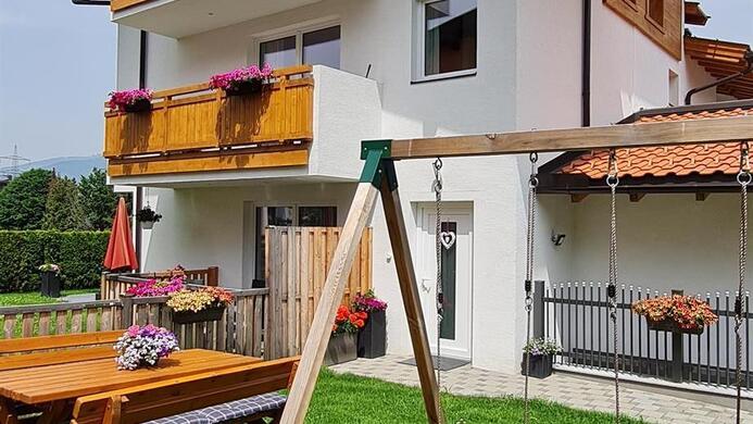A beautiful house with a balcony and a large garden. In the garden, there is a swing and a wooden seating area.
