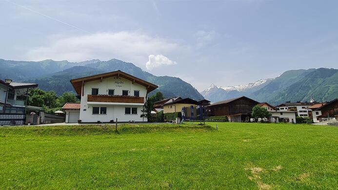 A peaceful landscape with green grass and several houses. In the background, majestic mountains and a blue sky can be seen.