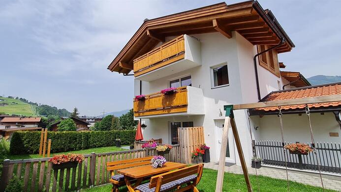 A cozy house with a wooden porch and balcony, surrounded by green grass. In the foreground, there is a wooden table with flowers.
