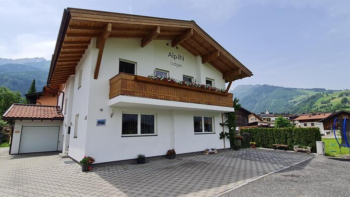 A modern two-story house with a wooden balcony. Gentle hills and mountains are visible in the background.