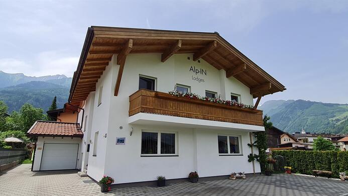 A modern house with wooden cladding and a balcony. In the background, green hills and mountains are visible.