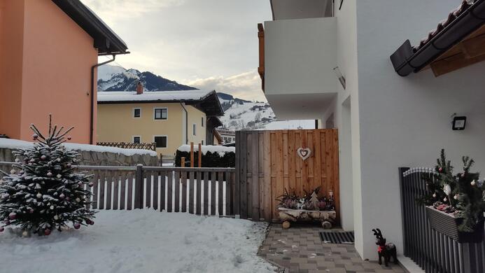 A conservatory with freshly fallen snow and a small Christmas tree. In the background, mountains and a few colorful houses are visible.