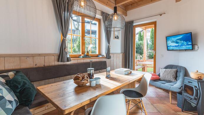 Ein gem&uuml;tlicher Wohnbereich mit einem Holztisch und modernen St&uuml;hlen. Gro&szlig;e Fenster lassen viel Tageslicht herein und bieten Blick ins Freie.