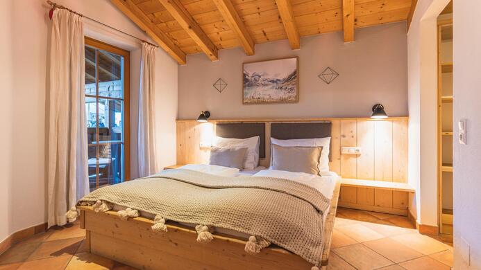 Ein gem&uuml;tliches Schlafzimmer mit einem gro&szlig;en Bett und Holzdecken. Es gibt ein Fenster mit Vorh&auml;ngen und warmem Licht, das den Raum einladend macht.