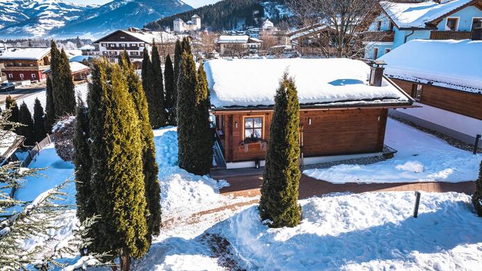 Eine malerische Winterlandschaft mit einem Holzhaus, umgeben von schneebedeckten B&auml;umen. Im Hintergrund sind Berge und ein blauer Himmel zu sehen.