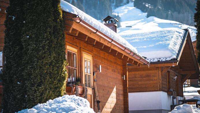 Ein idyllisches Holzhaus in den Bergen, umgeben von Schnee. Die Sonne scheint auf die schneebedeckten D&auml;cher und die malerische Landschaft.