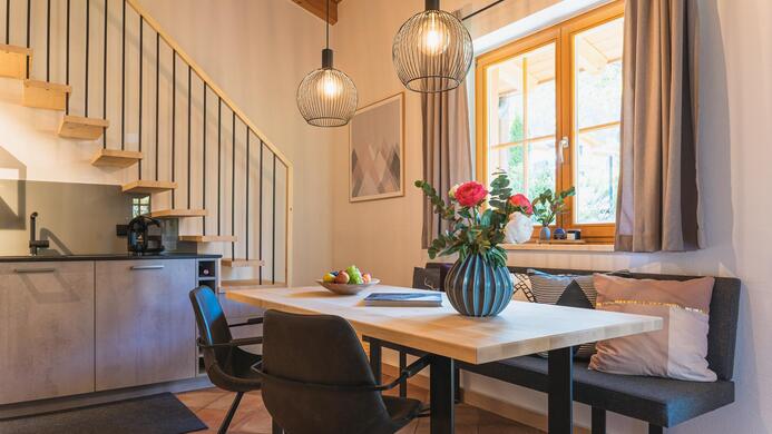 Ein gem&uuml;tlicher Essbereich mit einem Holztisch, modernen St&uuml;hlen und einer Vase mit Blumen. Der Raum hat gro&szlig;e Fenster und eine Treppe im Hintergrund.