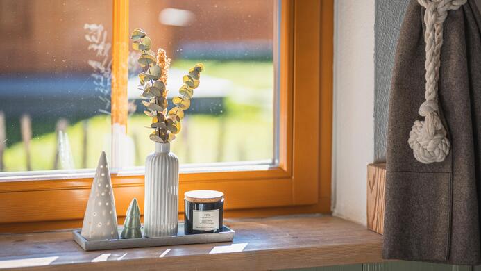 Ein Fensterbrett mit dekorativen Objekten wie einer Vase, Kerzen und kleinen Weihnachtsb&auml;umen. Die orangefarbene Fensterrahmung bringt warmes Licht in den Raum.