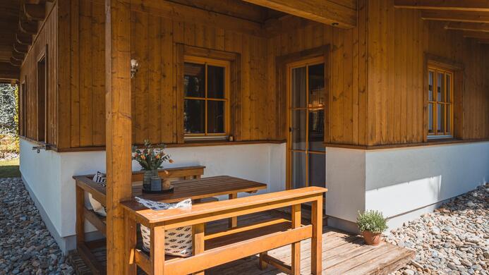 Eine einladende Holzh&uuml;tte mit einer gem&uuml;tlichen Terrasse. Auf dem Tisch steht eine Vase mit Blumen, und der Boden ist mit Kies bedeckt.