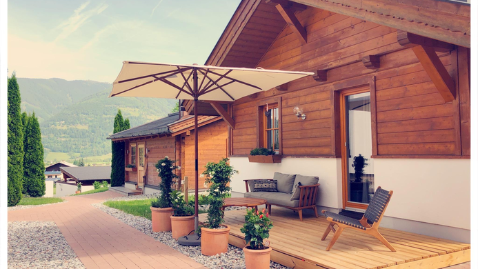 Ein gem&uuml;tliches Holzhaus mit einer Terrasse und einem Sonnenschirm. Vor dem Haus stehen Pflanzen in T&ouml;pfen und die Landschaft ist h&uuml;gelig mit Bergen im Hintergrund.