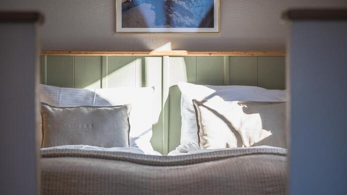 Ein gem&uuml;tliches Schlafzimmer mit einem Bett und hellen Kissen. Sanftes Licht f&auml;llt auf die Bettw&auml;sche und die Wand.