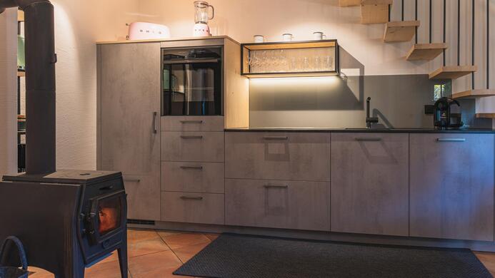 Eine moderne K&uuml;che mit grauen M&ouml;beln und Holzdetails. Im Vordergrund steht ein gem&uuml;tlicher Kaminofen.