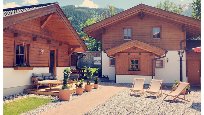 Eine malerische Holzhausanlage umgeben von gr&uuml;nen Bergen. Im Innenhof stehen Liegen und Pflanzen, die eine einladende Atmosph&auml;re schaffen.