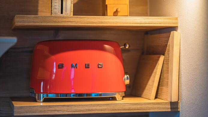 Ein roter Toaster von SMEG steht auf einem Holzregal. Darunter sind einige B&uuml;cher und ein kleiner Holzgegenstand zu sehen.