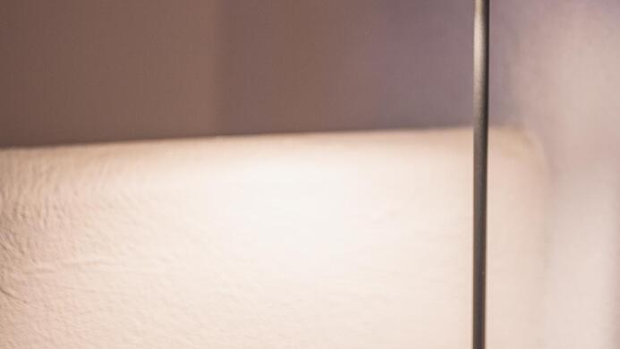 Eine moderne Stehlampe steht neben einem grauen Sessel. Der Sessel hat ein orangefarbenes Kissen und der Hintergrund ist hell beleuchtet.