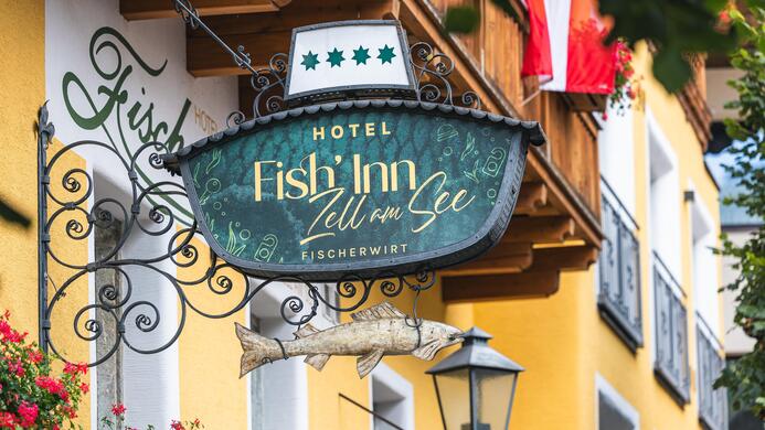 Ein einladendes Schild des Hotels "Fish Inn" in Zell am See. Das designte Schild zeigt einen Fisch und ist von Blumen umgeben.