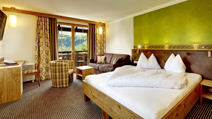 Ein modernes Hotelzimmer mit einem Doppelbett, einer gem&uuml;tlichen Sitzecke und einem Balkon. Die Einrichtung kombiniert Holz und farbige Akzente, sodass eine einladende Atmosph&auml;re entsteht.