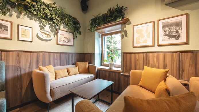 Ein gem&uuml;tlicher Raum mit beigen Sofas und einem Holztisch. An den W&auml;nden h&auml;ngen Bilder und Pflanzen sorgen f&uuml;r eine angenehme Atmosph&auml;re.