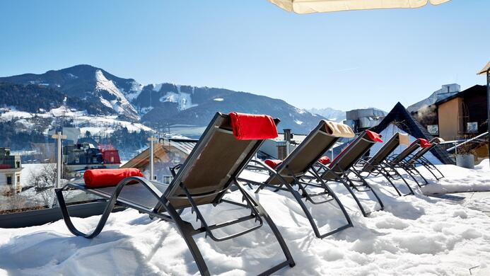 Liegest&uuml;hle stehen im Schnee und bieten einen Blick auf die schneebedeckten Berge. Der Himmel ist klar und sonnig.