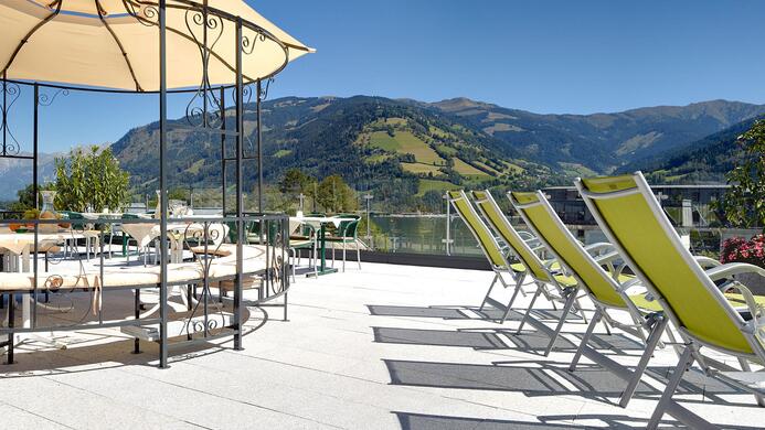 Eine sonnige Terrasse mit Liegen und einem Sonnenschirm. Im Hintergrund sind gr&uuml;ne Berge und ein blauer Himmel zu sehen.