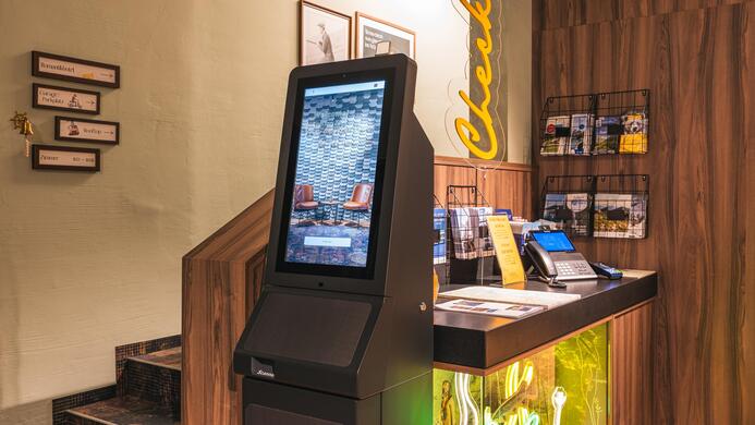 Ein moderner Check-in-Automat steht an einem Empfangstresen. Im Hintergrund sind dekorative Elemente und Informationsmaterialien sichtbar.