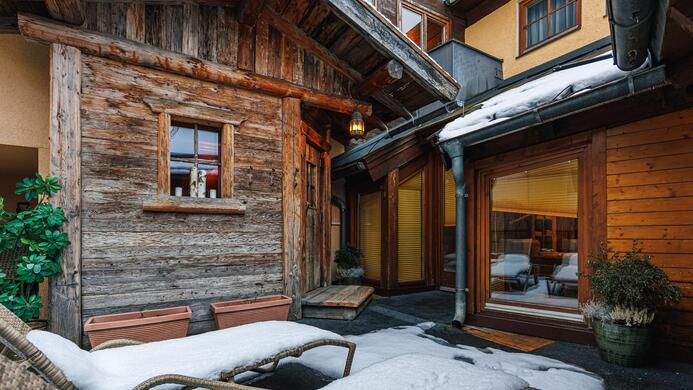 Ein gem&uuml;tlicher Innenhof mit Holzstruktur und Schneebedeckung. Sicht auf bequeme Liegen und gro&szlig;e Fenster des angrenzenden Raumes.