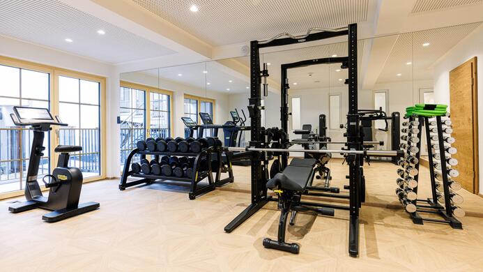 Ein modernes Fitnessstudio mit verschiedenen Trainingsger&auml;ten und freien Gewichten. Der Raum ist hell und einladend, mit gro&szlig;en Fenstern und Holzfu&szlig;boden.