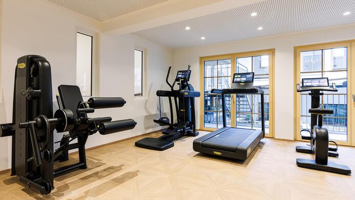 Ein modernes Fitnessstudio mit verschiedenen Trainingsger&auml;ten. Der Raum ist hell erleuchtet und hat gro&szlig;e Fenster.