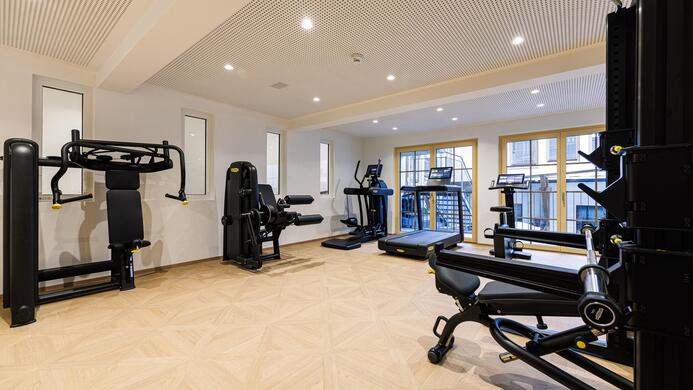 Ein moderner Fitnessraum mit verschiedenen Trainingsger&auml;ten. Der Raum hat gro&szlig;e Fenster, die viel Tageslicht hereinlassen.