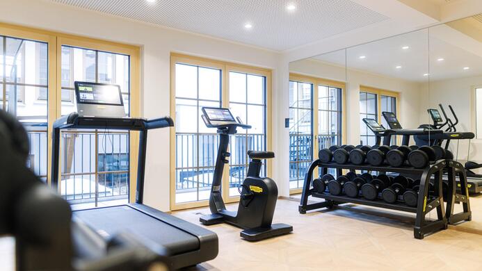 Ein modernes Fitnessstudio mit Laufb&auml;ndern, Gewichten und gro&szlig;en Fenstern. Der Raum ist hell und einladend.