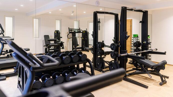 Ein modernes Fitnessstudio mit verschiedenen Trainingsger&auml;ten. Es gibt Hanteln, eine Bank und Spiegel an den W&auml;nden.