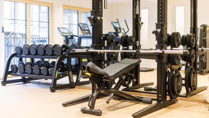 Ein modernes Fitnessstudio mit Hanteln und einer Gewichtsbank. Im Hintergrund sind weitere Fitnessger&auml;te sichtbar.