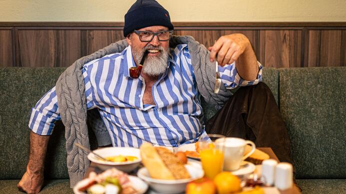 Ein Mann mit Bart sitzt auf einer Couch und genie&szlig;t sein Fr&uuml;hst&uuml;ck. Auf dem Tisch sind verschiedene Speisen und Getr&auml;nke, darunter Brot und Obst.