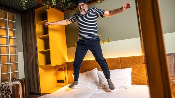 Ein Mann springt fr&ouml;hlich auf einem Bett in einem modernen Zimmer. Die Atmosph&auml;re ist lebhaft und einladend.