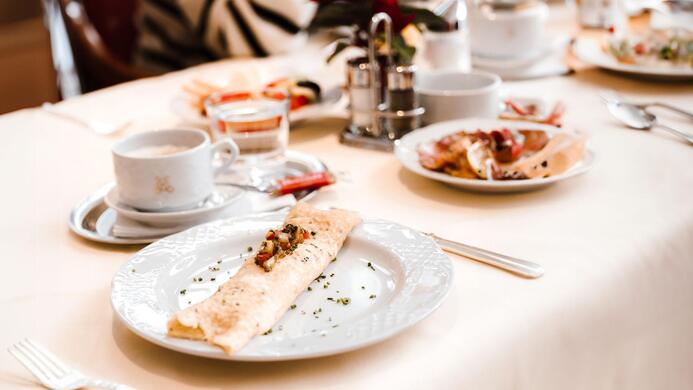 An elegant breakfast table with various dishes. In the foreground, there is a plate with a crepe and a cup of coffee.